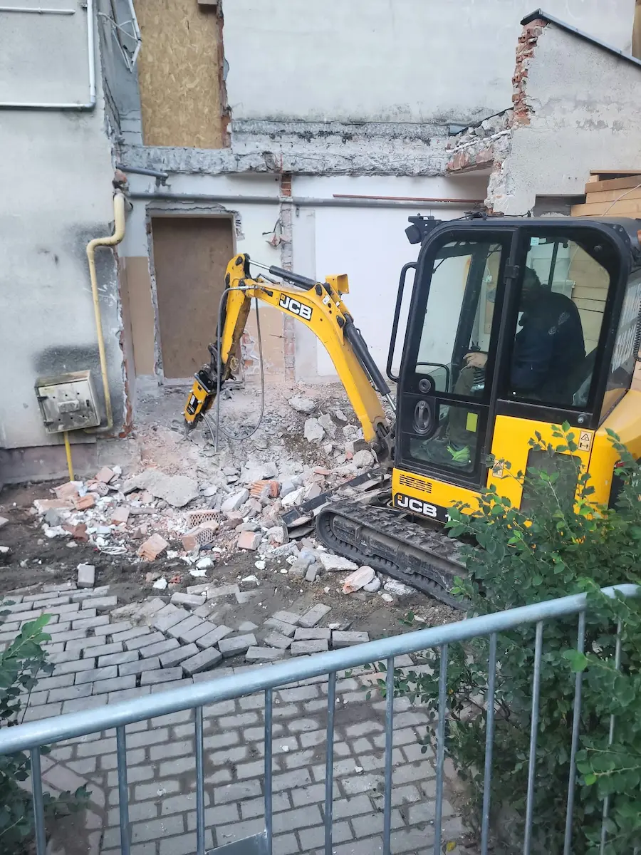 Rozbiórka budynku w Siedlcach mini koparką JCB — operator i pracownik przy ścianie, gruz na terenie, ogrodzenie ochronne, prace rozbiórkowe Ziemne.net